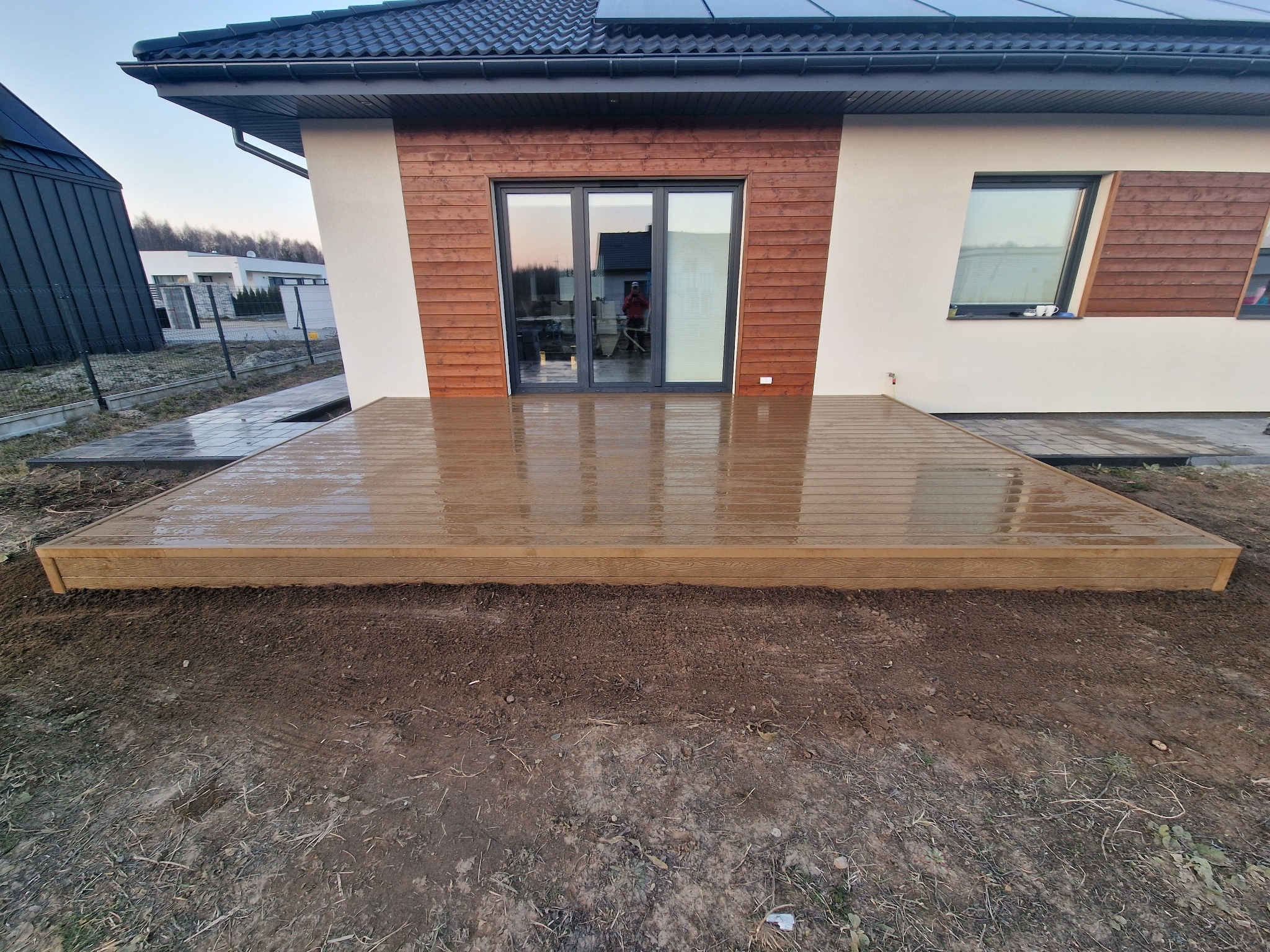 Nowoczesny, drewniany taras przydomowy z widocznymi refleksami wody po deszczu, z brązową elewacją i oknami domu w tle.