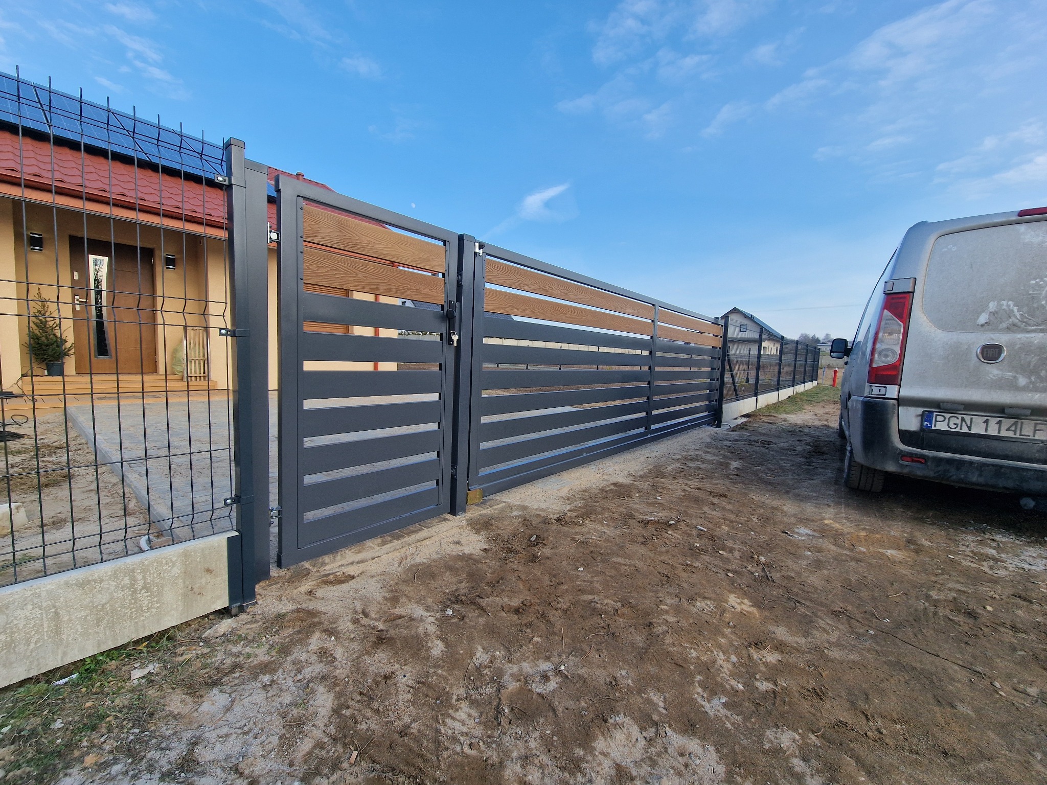 Nowoczesne ogrodzenie panelowe z drewnianymi akcentami, brama otwarta. Widoczny dom z panelami słonecznymi i auto dostawcze. Słoneczny dzień.