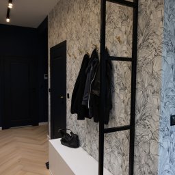 P.P.H.U Intlux - Nowoczesny przedpokój z tapetą w tropikalne liście, czarnymi drzwiami i złotymi wieszakami, uzupełniony białą szafką i czarną torebką.