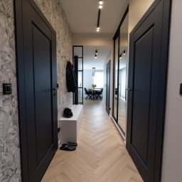 P.P.H.U Intlux - Nowoczesny przedpokój z parkietem w jodełkę, czarnymi drzwiami i szafą z lustrem, oświetleniem szynowym i tapetą w motywy roślinne.