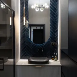 Nowoczesna łazienka z owalnym lustrem obramowanym granatowymi płytkami ułożonymi w jodełkę, czarną umywalką nablatową i szafką z ryflowanymi frontami.