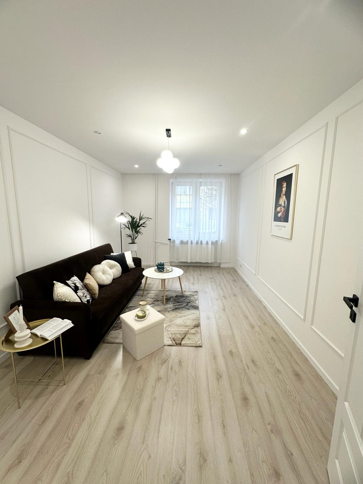 Jasne wnętrze salonu po remoncie: panele podłogowe w odcieniach drewna, ciemna sofa z dekoracyjnymi poduszkami, białe ściany z dekoracyjnymi listwami, okno z firanką i minimalistyczne oświetlenie.