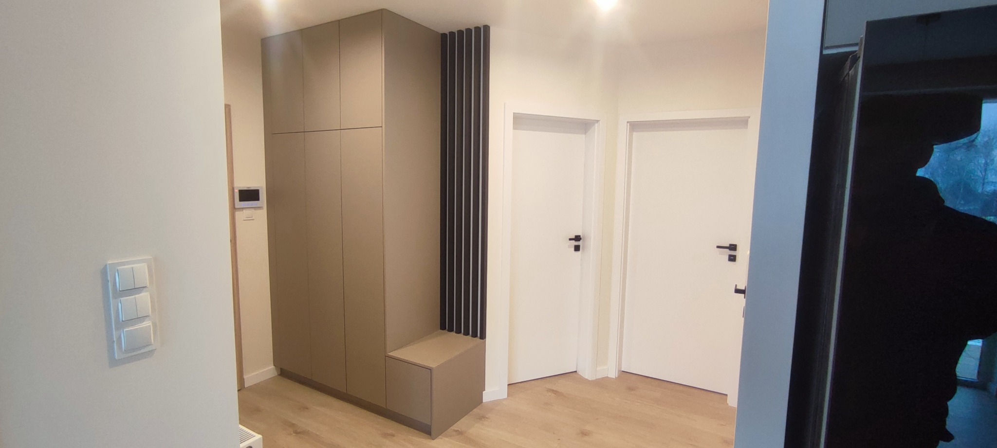 Nowoczesny przedpokój z zabudowaną szafą w kolorze taupe z siedziskiem i dekoracyjnymi czarnymi listwami, białe drzwi z czarnymi klamkami, minimalistyczne oświetlenie.