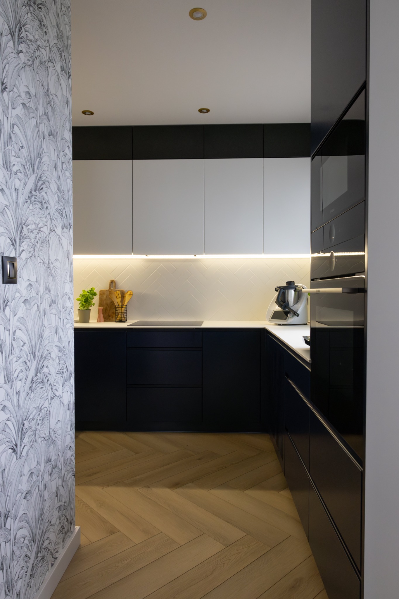 Nowoczesna kuchnia z czarno-białymi szafkami, podświetleniem LED, parkietem w jodełkę i tapetą w liście, widziana z perspektywy wejścia.