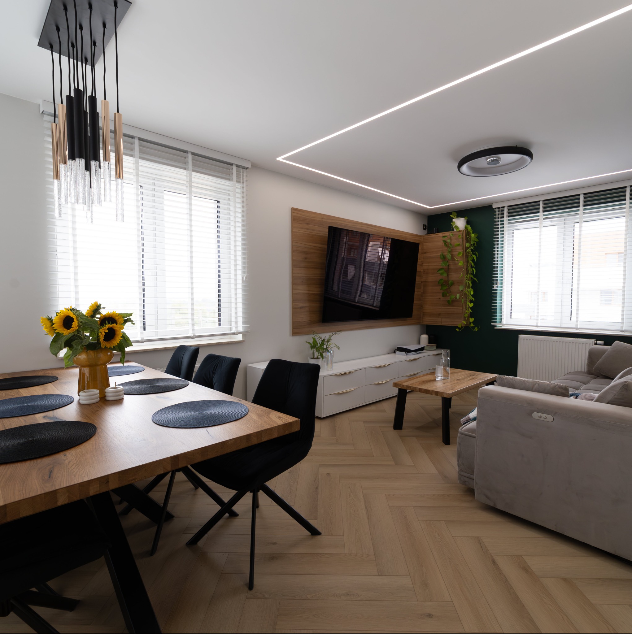 Nowoczesne wnętrze salonu z jadalnią: drewniany stół z czarnymi krzesłami, parkiet ułożony w jodełkę, szary narożnik, ściana z telewizorem, oświetlenie LED na suficie i bukiet słoneczników na stole.