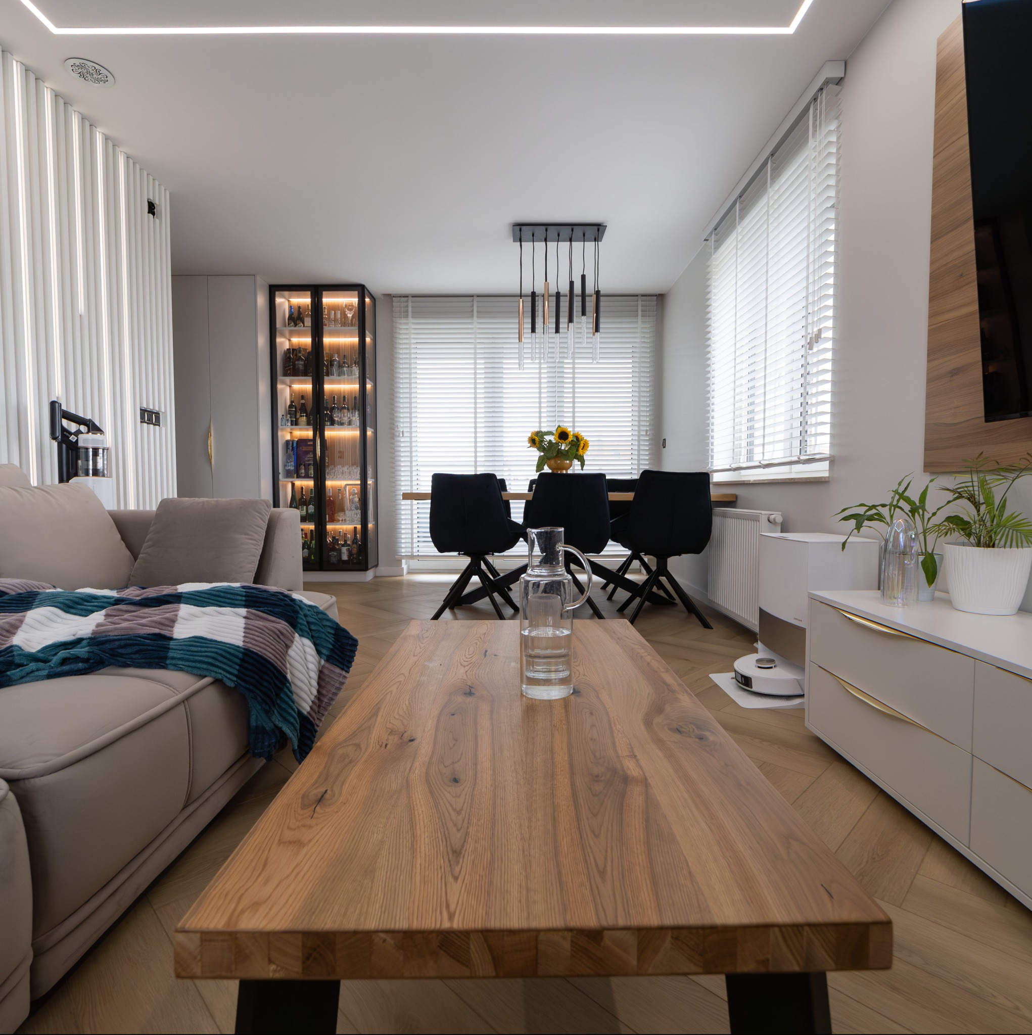 Jasne, nowocześnie urządzone wnętrze salonu z jadalnią, z drewnianym stołem i krzesłami, szklaną witryną z oświetleniem, parkietem w jodełkę i robotem sprzątającym.