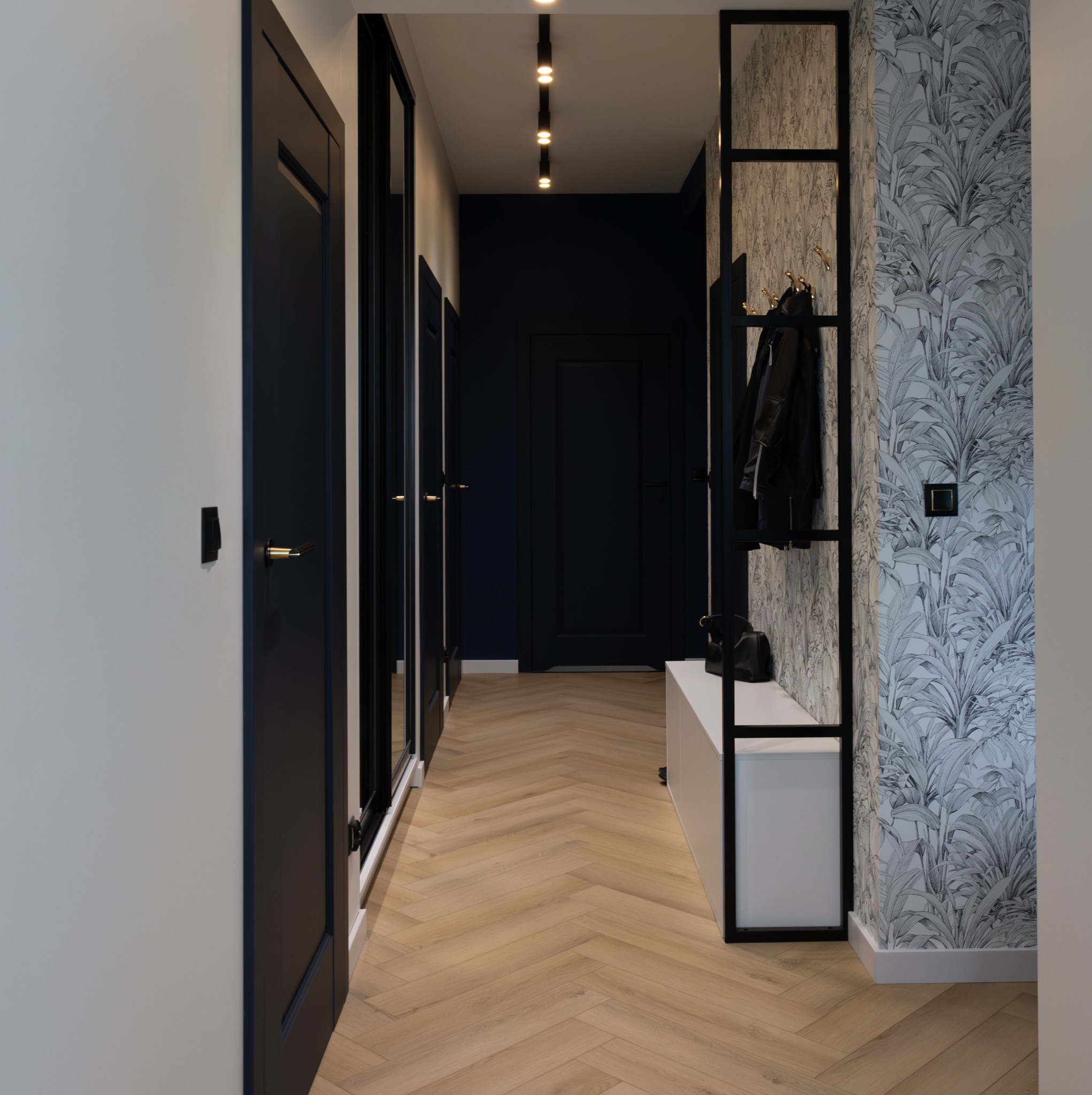 Nowoczesny przedpokój z parkietem w jodełkę, ciemnymi drzwiami i szafami w zabudowie, tapetą w roślinny wzór oraz czarnym regałem z wieszakami i białą ławką.