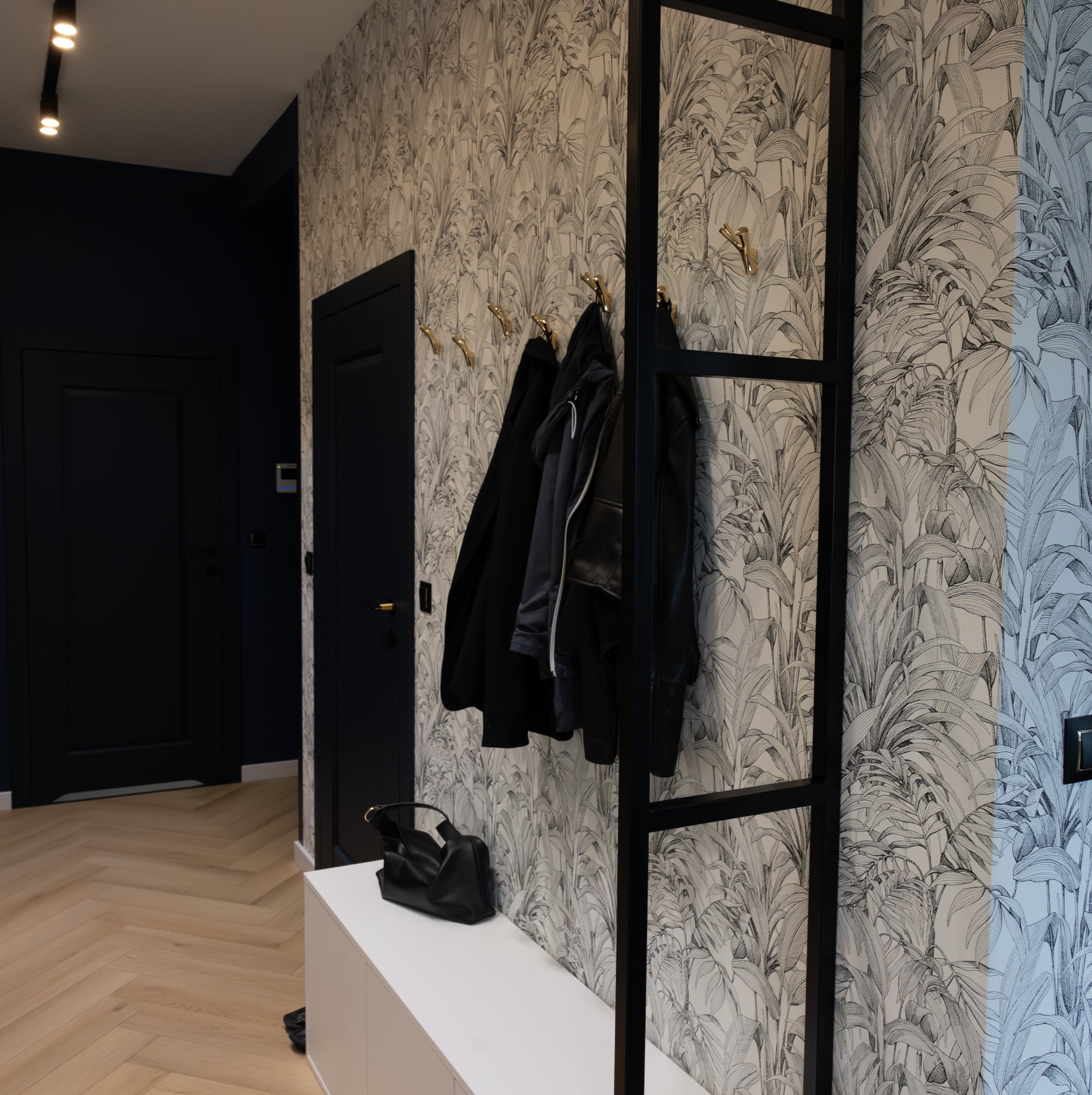 Nowoczesny przedpokój z tapetą w tropikalne liście, czarnymi drzwiami i złotymi wieszakami, uzupełniony białą szafką i czarną torebką.