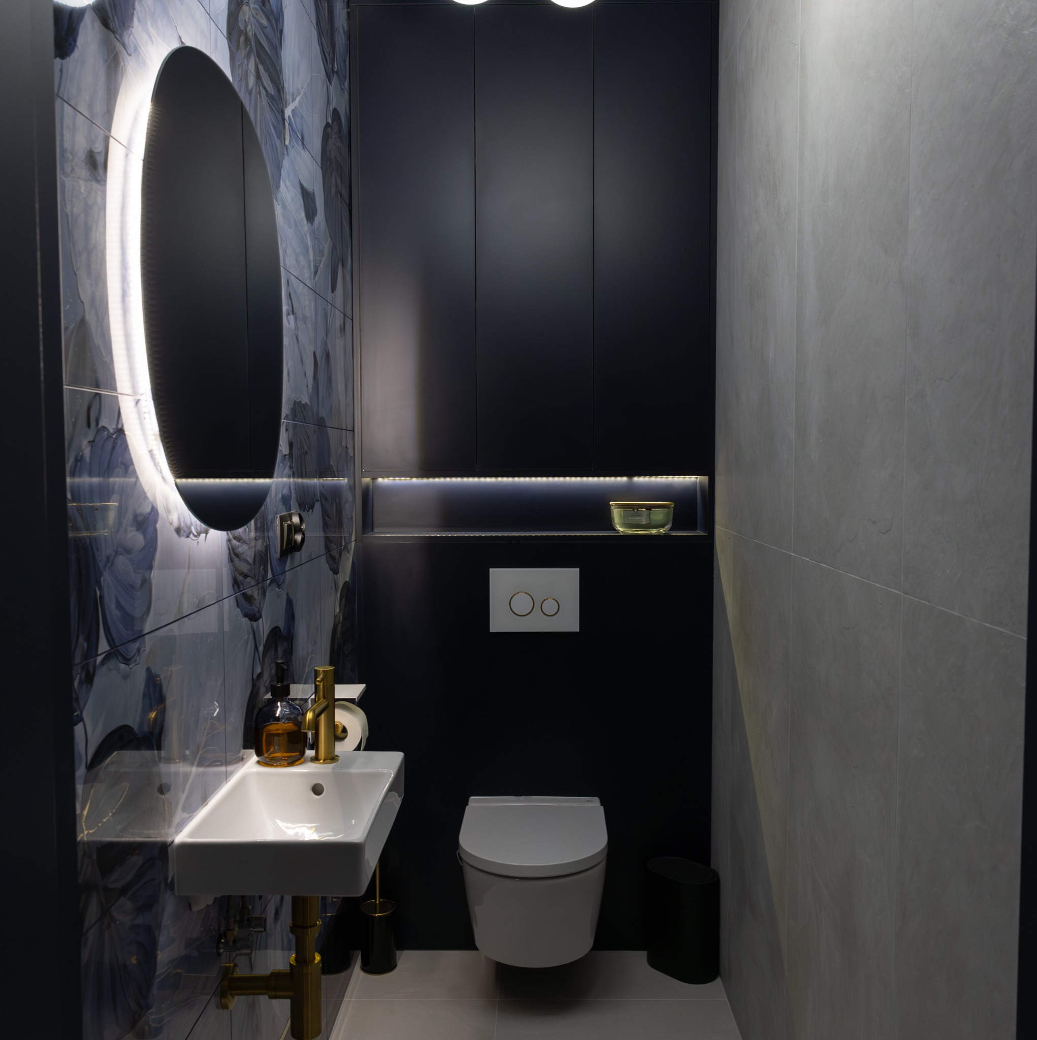 Nowoczesna łazienka w odcieniach szarości i granatu z umywalką na złotym stelażu, podświetlanym owalnym lustrem i wiszącą toaletą.