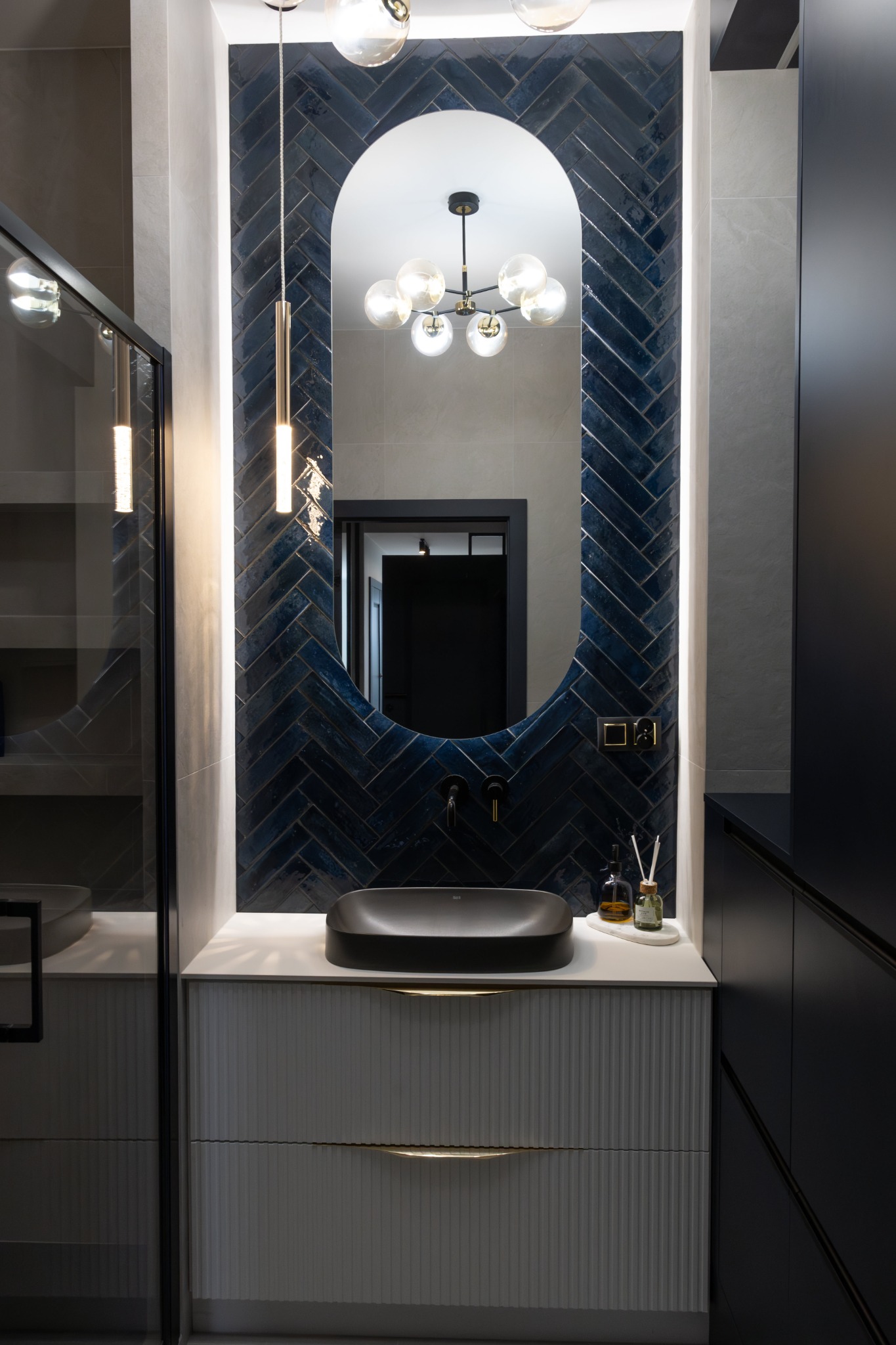 Nowoczesna łazienka z owalnym lustrem obramowanym granatowymi płytkami ułożonymi w jodełkę, czarną umywalką nablatową i szafką z ryflowanymi frontami.