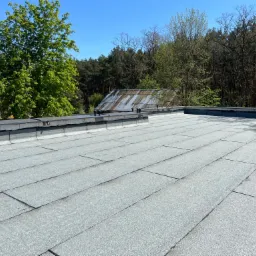 Płaski dach pokryty szarymi panelami bitumicznymi, widok z góry na tle zieleni drzew i błękitnego nieba.