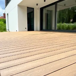 Nowoczesny taras z desek kompozytowych, przylegający do białego budynku z dużymi przesuwnymi oknami, widok z niskiej perspektywy ukazujący strukturę desek i otoczenie domu.