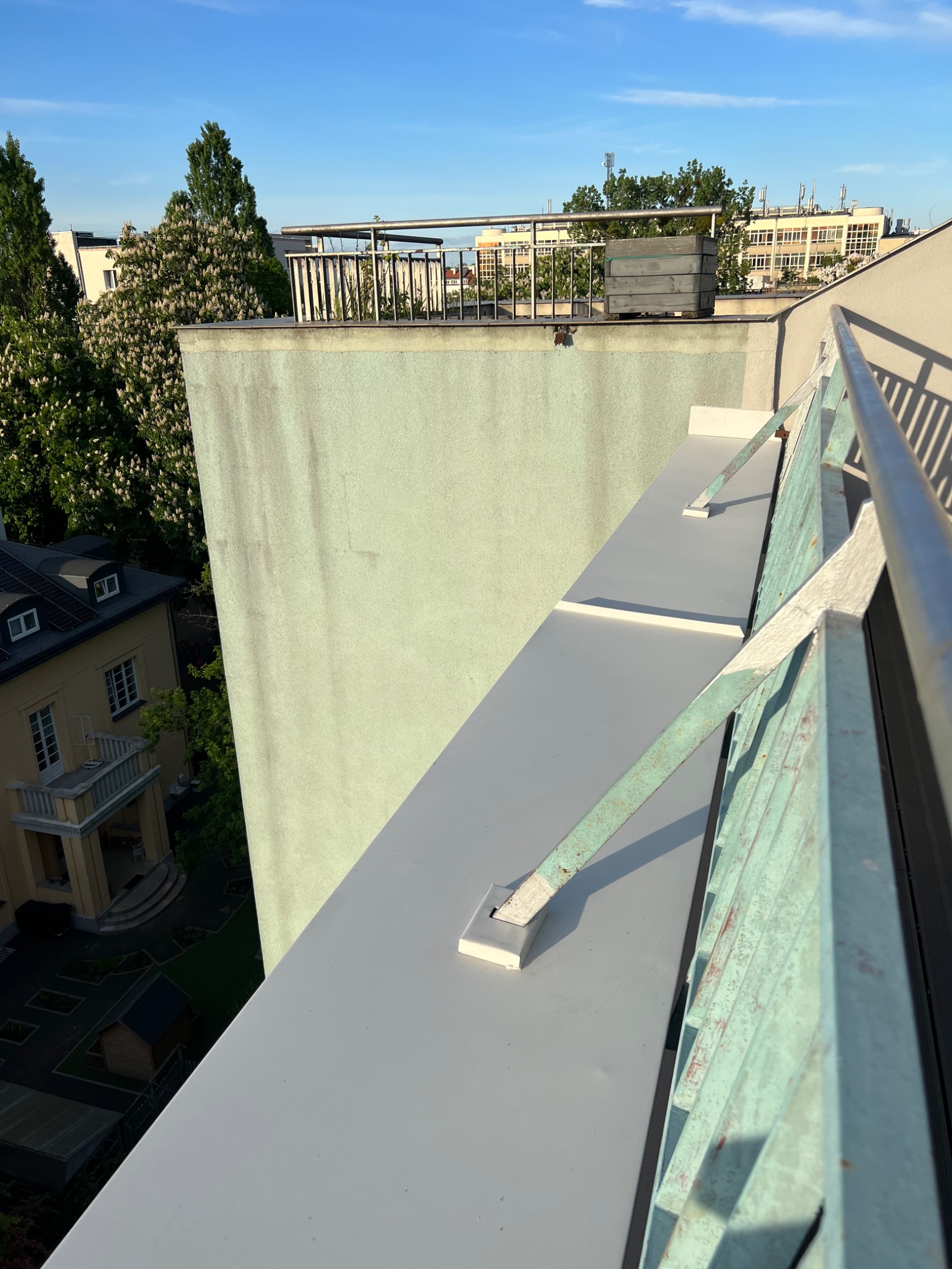 Widok z góry na płaski dach budynku z szarą, gładką powierzchnią, z zielonkawą ścianą budynku w tle i fragmentem metalowej balustrady w kolorze morskim, z widokiem na okoliczną zabudowę i drzewa.