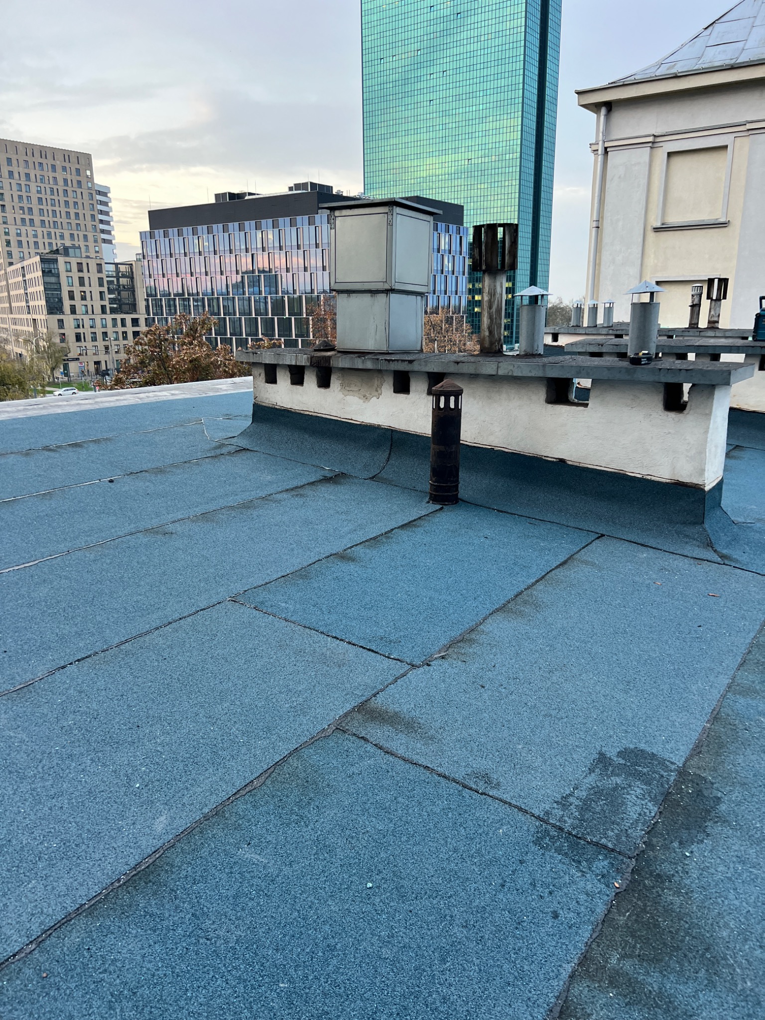 Niebieski dach pokryty papą termozgrzewalną z kominami i elementami wentylacyjnymi na tle miejskiego krajobrazu z wieżowcami.