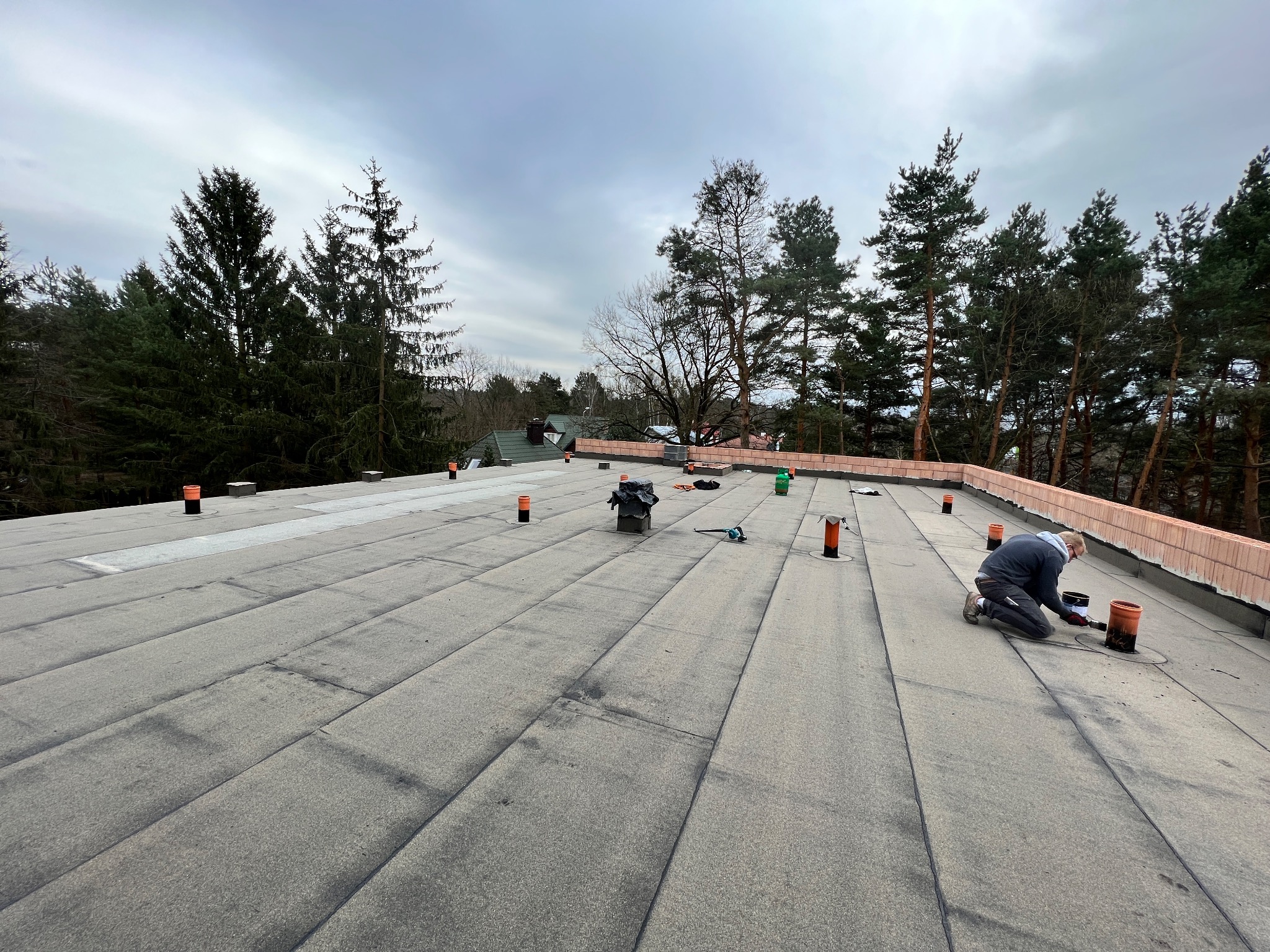 Pracownik na kolanach uszczelnia kominek wentylacyjny na płaskim dachu pokrytym papą, widoczne narzędzia i materiały, w tle drzewa i pochmurne niebo.