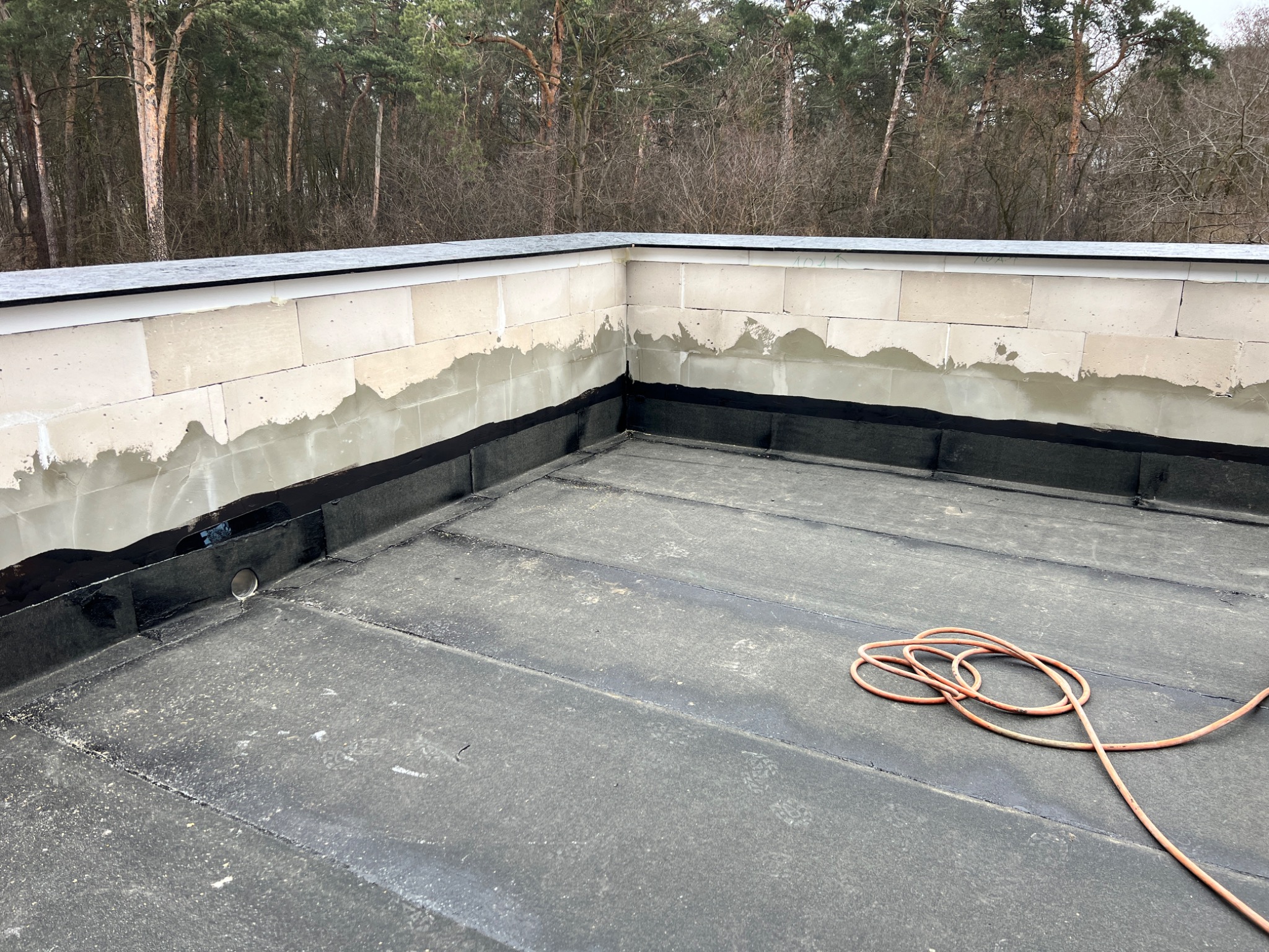 Płaski dach pokryty papą, z widocznymi fragmentami muru z białych bloczków i czarną hydroizolacją przy krawędziach; w tle drzewa.