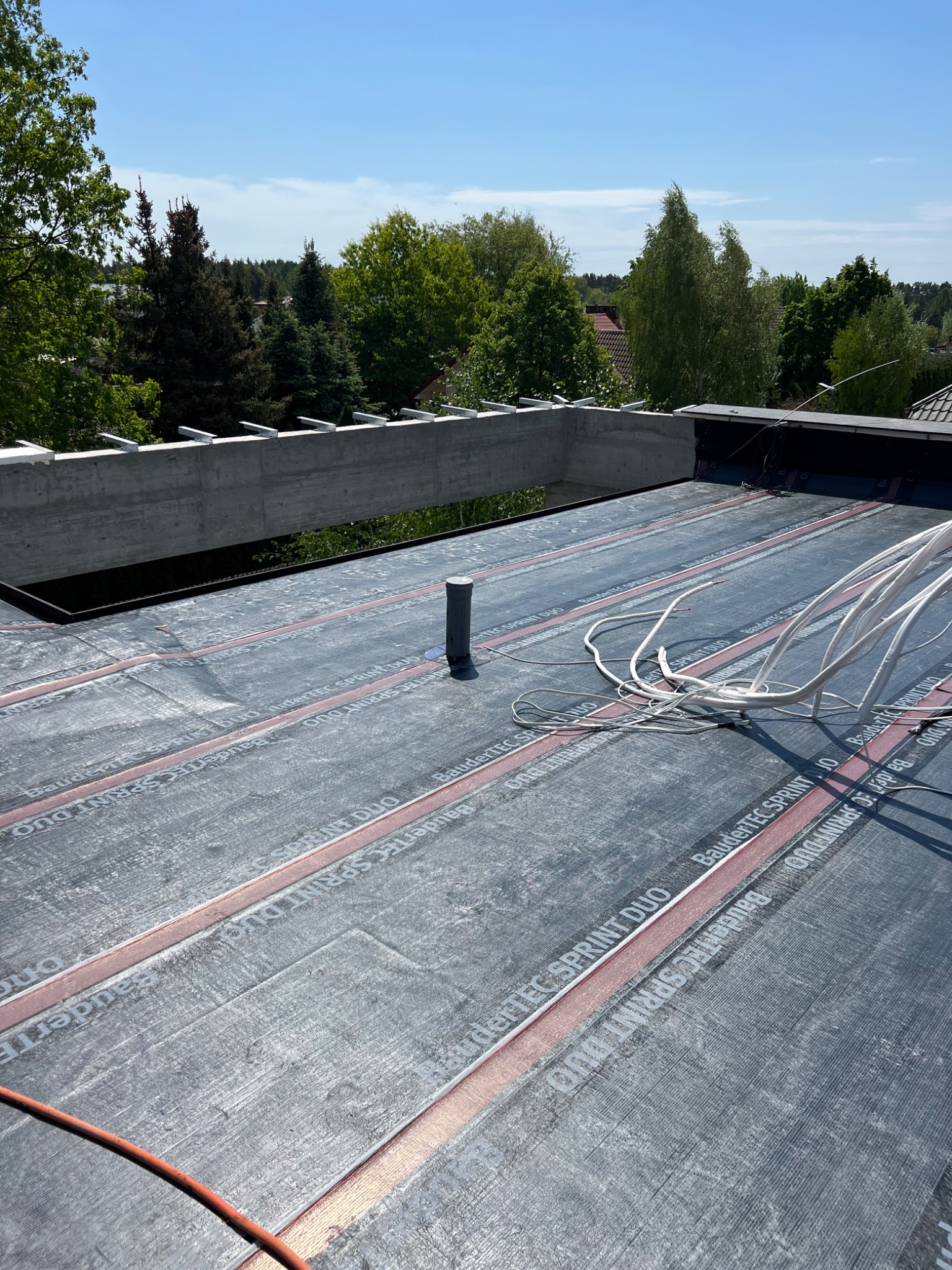 Czarny dach płaski z papy termozgrzewalnej BauderTEC SPRINT DUO z widocznymi pasami zgrzewu, wystającą rurką odpowietrznika i przewodami elektrycznymi, betonowy element konstrukcyjny dachu w tle.