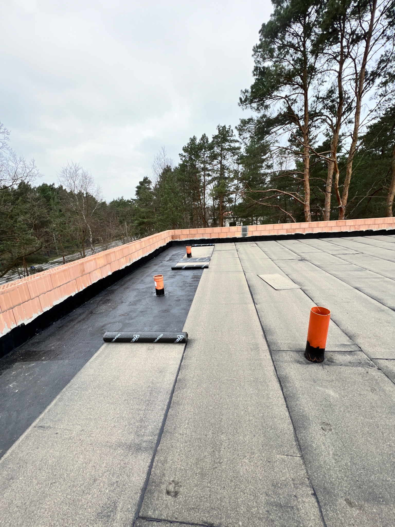 Hydroizolacja płaskiego dachu z użyciem papy termozgrzewalnej, widoczne kominki wentylacyjne i ceglana attyka w tle drzewa.