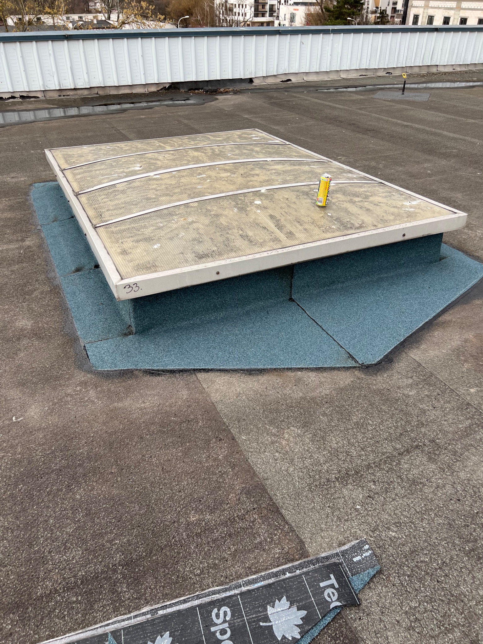 Płaski dach z włazem dachowym oświetlającym wnętrze, zabezpieczonym blachą i papą, z widoczną puszką po napoju na powierzchni włazu i resztkami materiałów dekarskich w rogu.