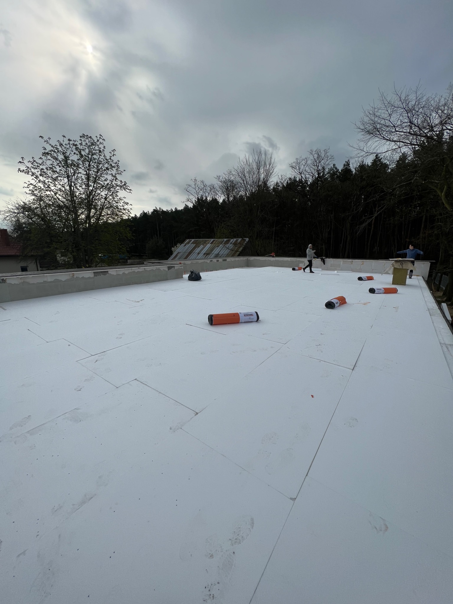 Montaż białych płyt izolacyjnych na dachu budynku w trakcie prac, widoczne rolki materiału i dwie osoby pracujące na tle pochmurnego nieba i drzew.