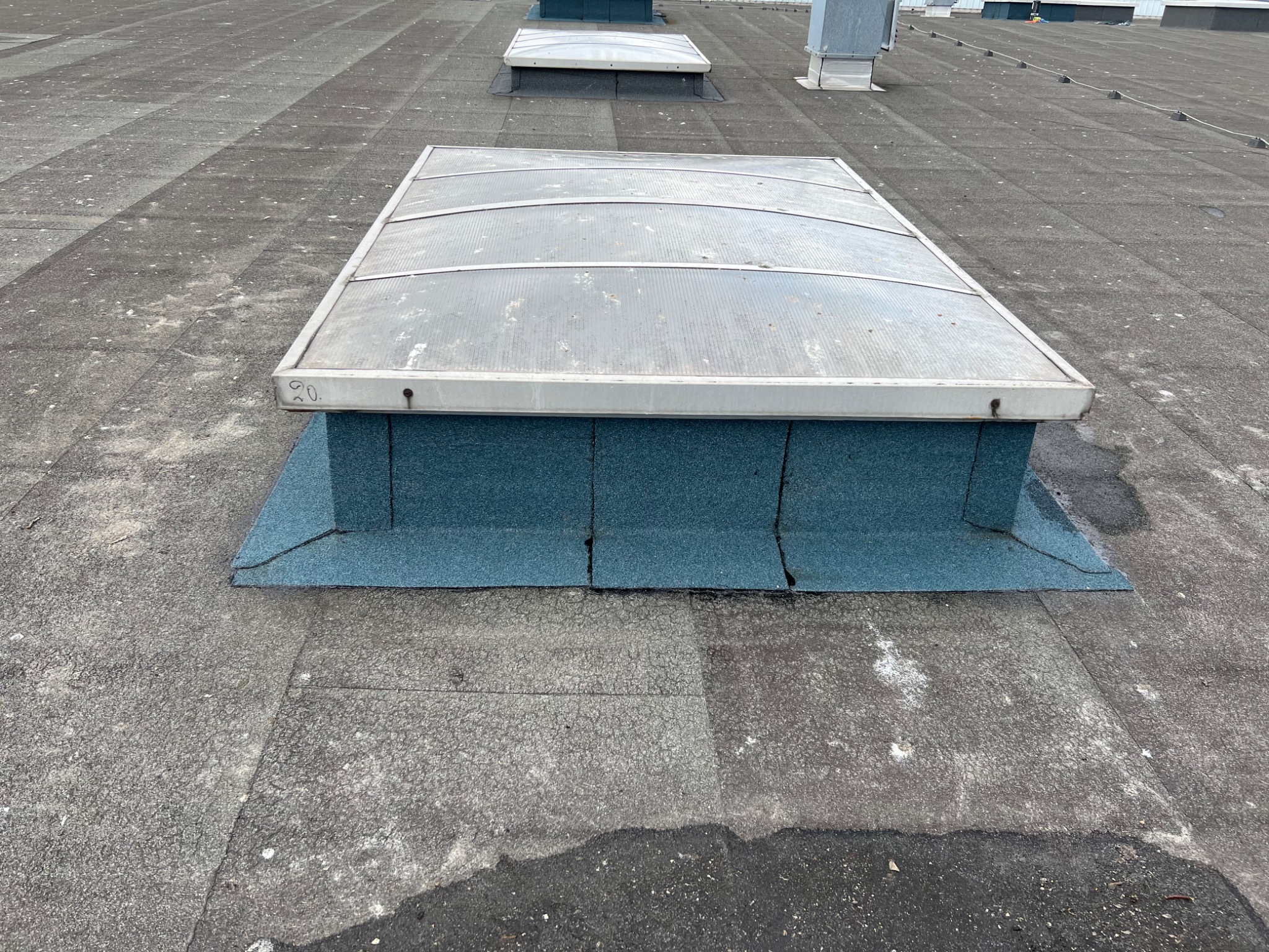 Płaski dach pokryty papą z świetlikiem dachowym o przezroczystej, lekko zabrudzonej powierzchni, otoczonym niebieską obróbką blacharską. W tle widoczne inne świetliki i elementy wentylacji.