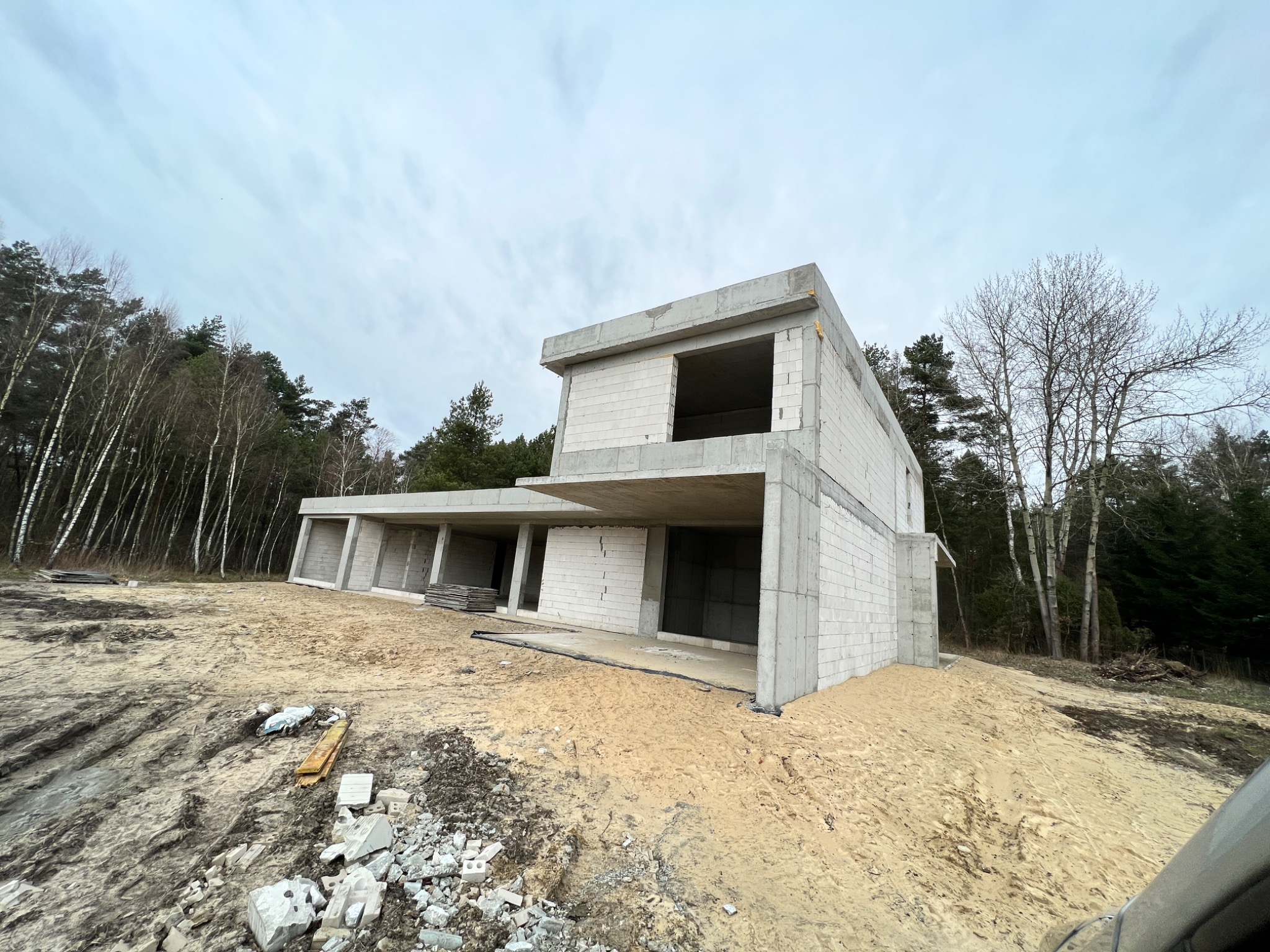 Surowy stan dwukondygnacyjnego domu z betonu komórkowego w trakcie budowy, widok z dołu na tle lasu i pochmurnego nieba.