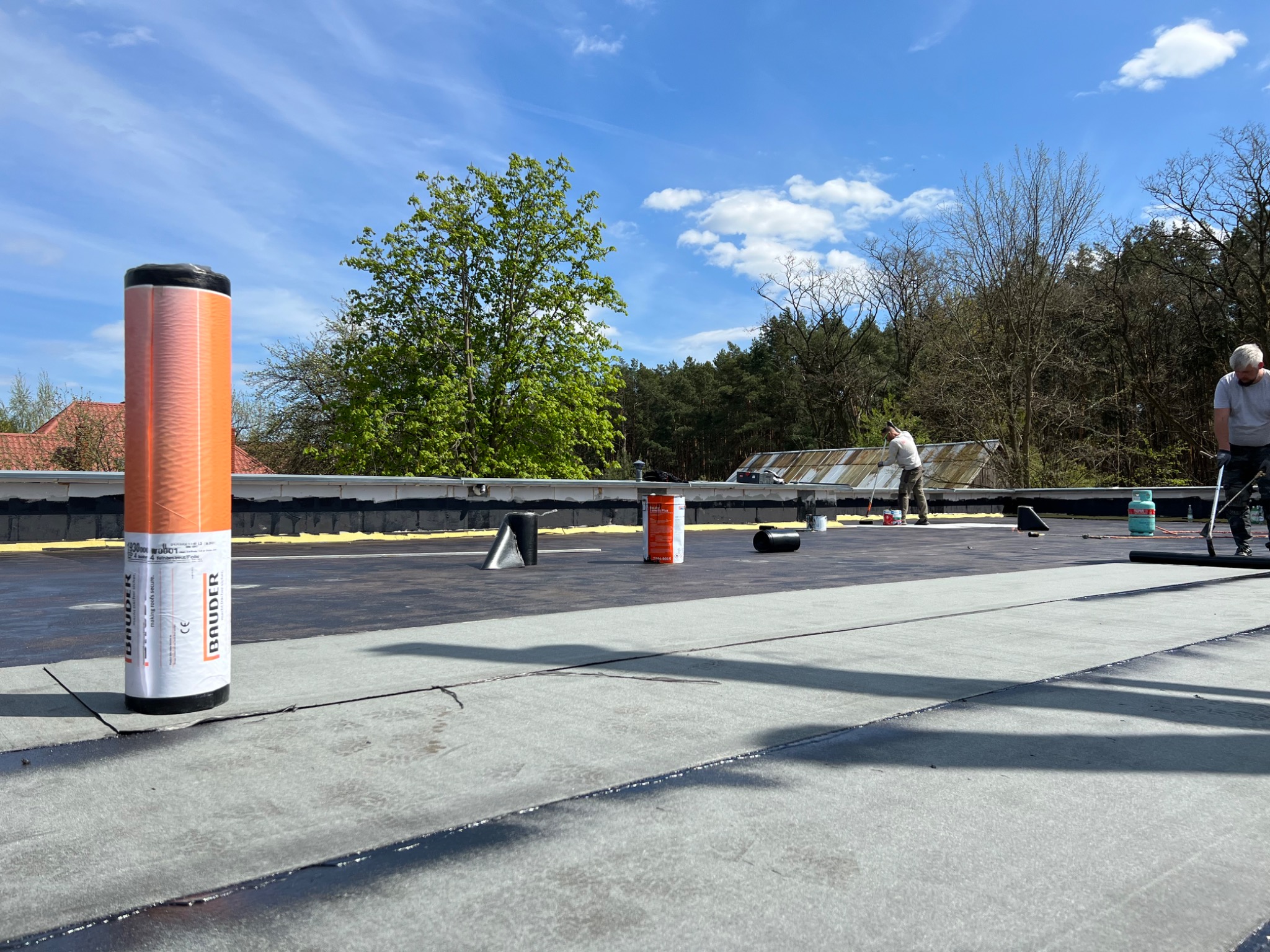 Ekipa dekarzy podczas prac na płaskim dachu pokrytym papą, widoczny duży rulon papy, butla z gazem i narzędzia dekarskie, w tle zieleń drzew i błękitne niebo.