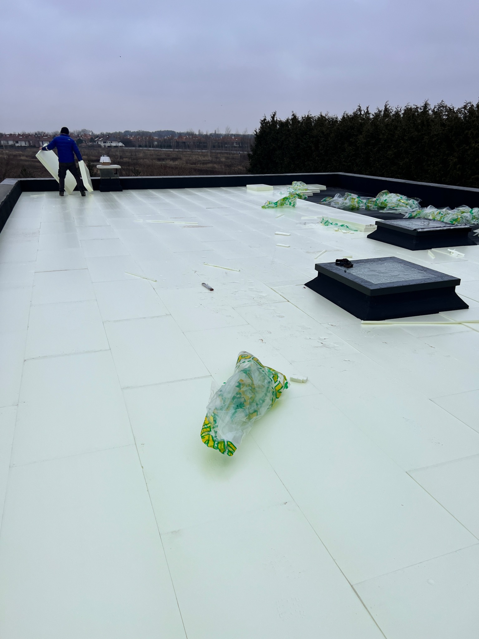 Prace na dachu płaskim: białe płyty izolacyjne układane przez pracownika, z widocznymi świetlikami dachowymi i resztkami opakowań po izolacji.