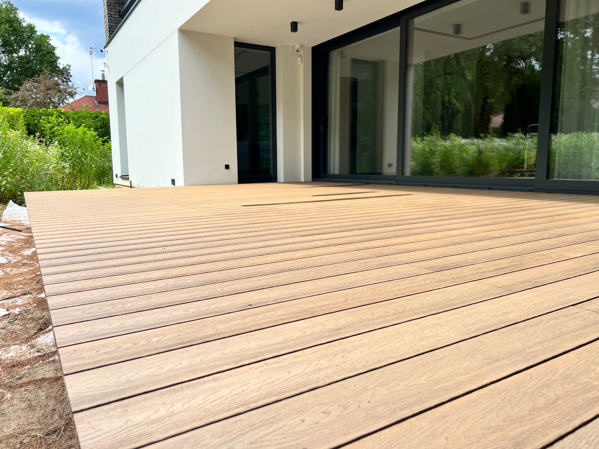 Nowoczesny taras z desek kompozytowych, przylegający do białego budynku z dużymi przesuwnymi oknami, widok z niskiej perspektywy ukazujący strukturę desek i otoczenie domu.