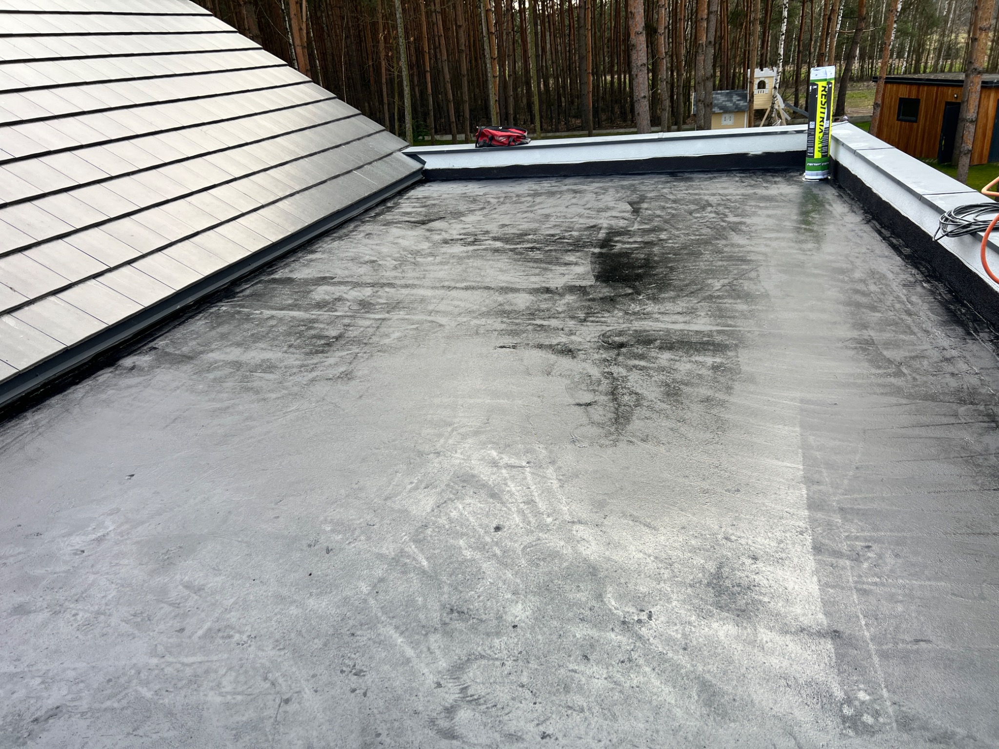 Świeżo zaizolowany, płaski dach pokryty czarną membraną bitumiczną, widoczne narzędzia i materiały montażowe, w tle las iglasty i zabudowania gospodarcze.