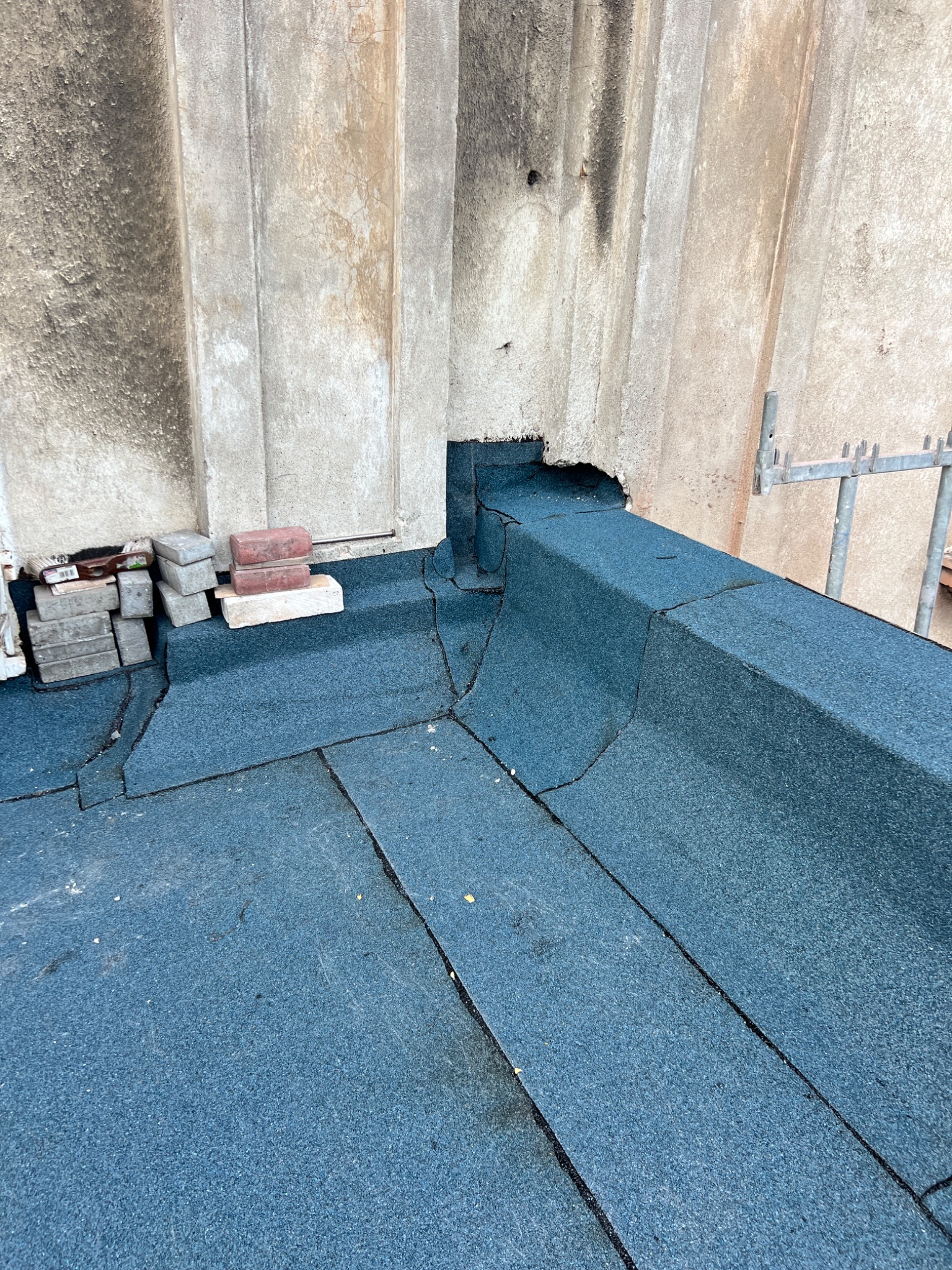 Niebieski dach pokryty papą z widocznymi pęknięciami i łatami. Na dachu leżą cegły i bloczki betonowe. W tle szara, brudna ściana z widocznym otworem i fragment rusztowania.