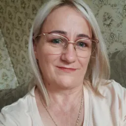 Portret uśmiechniętej kobiety z blond włosami w okularach z czerwoną oprawką, na szyi złoty łańcuszek, w tle tapeta w kwiaty i fragment sofy.