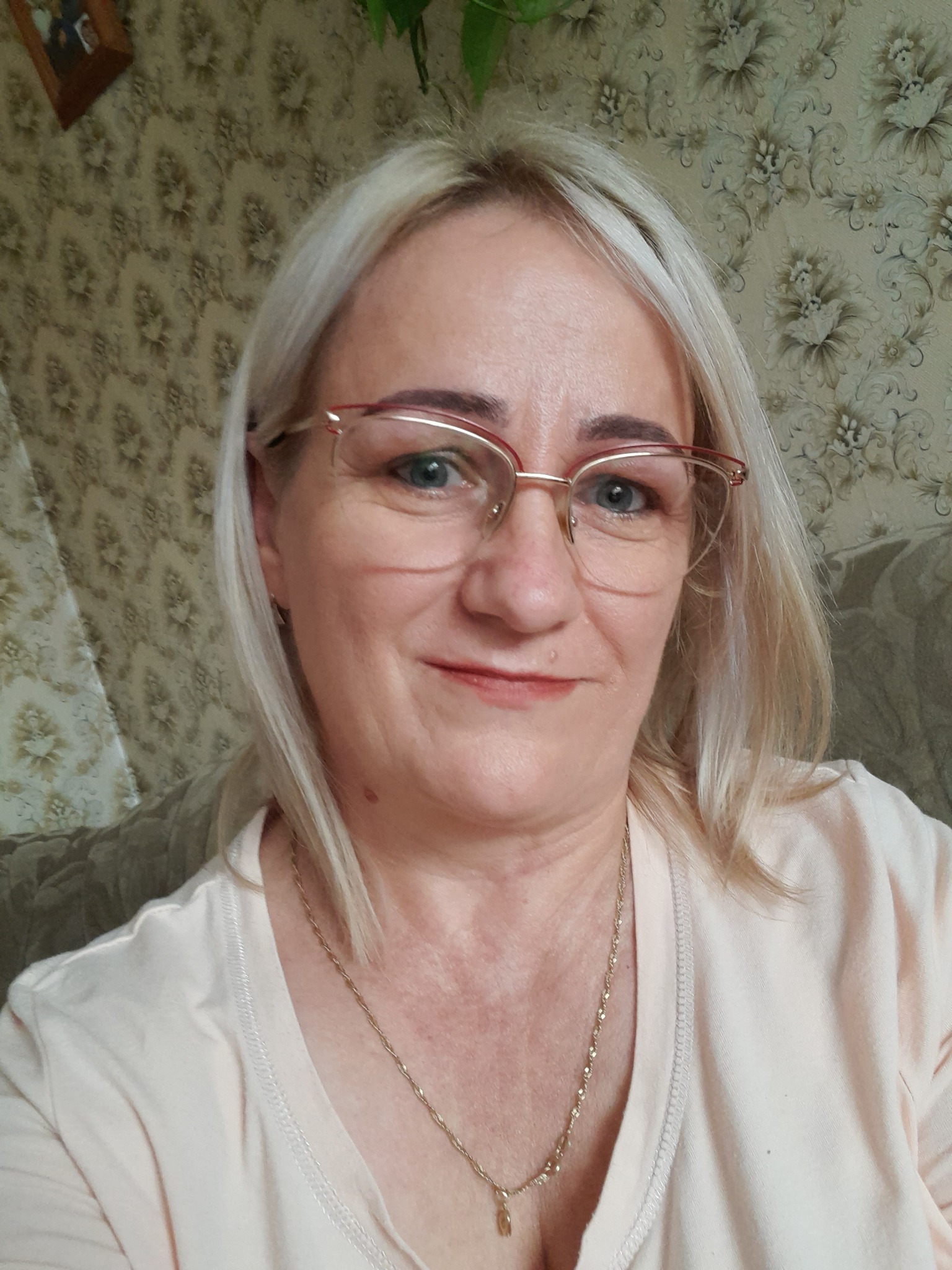 Portret uśmiechniętej kobiety z blond włosami w okularach z czerwoną oprawką, na szyi złoty łańcuszek, w tle tapeta w kwiaty i fragment sofy.