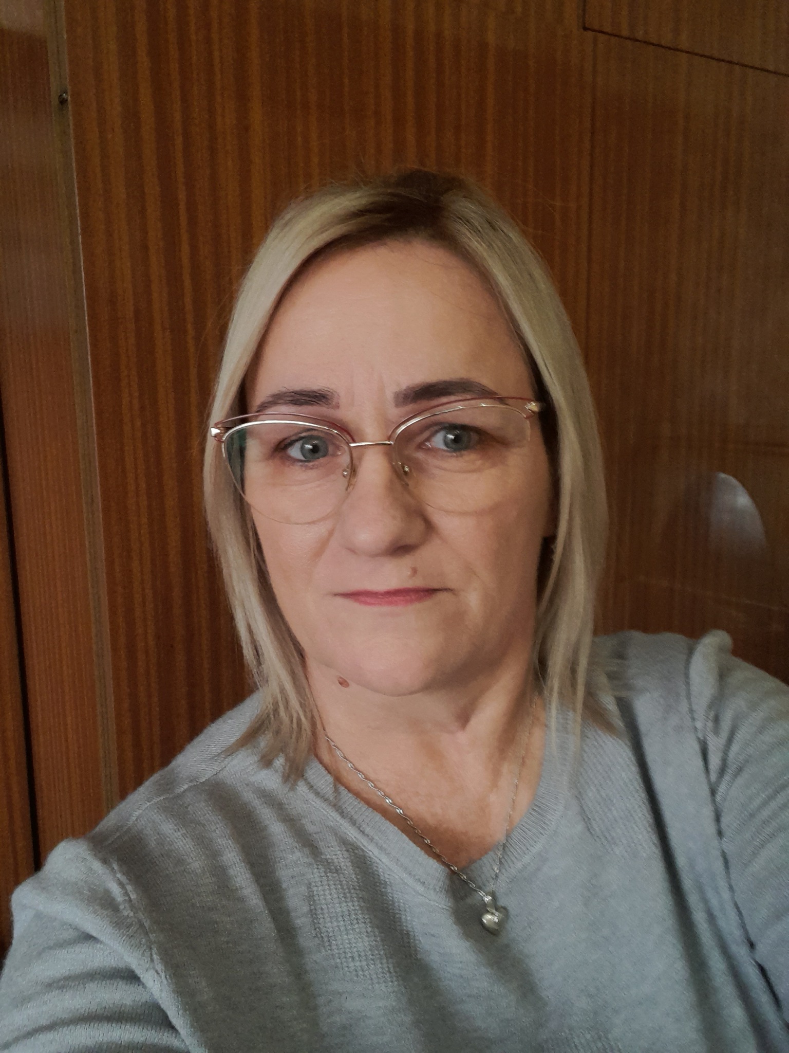 Portret kobiety w okularach, z blond włosami i srebrnym naszyjnikiem w kształcie serca, na tle drewnianej ściany.