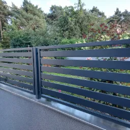 Nowoczesne, poziome przęsła ogrodzenia z metalu w kolorze grafitowym, zamontowane na niskim murku, z widokiem na bujną zieleń ogrodu z drzewami i krzewami.