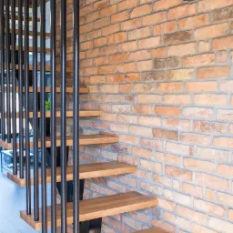 Drewniane schody wspornikowe z czarną, pionową balustradą na tle ceglanej ściany w stylu loftowym.