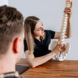 Kobieta w ciemnej koszulce prezentuje model kręgosłupa pacjentowi siedzącemu przy drewnianym stole, na którym leży laptop.
