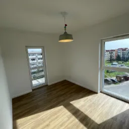 Pusty pokój w Łodzi po remoncie: świeżo położone panele podłogowe w odcieniach brązu, białe ściany, okno z widokiem na osiedle i nowoczesna lampa sufitowa.