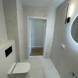 Nowoczesna łazienka z białymi płytkami imitującymi marmur, podwieszaną toaletą z czarnym przyciskiem, okrągłym lustrem z podświetleniem LED i minimalistycznym oświetleniem ściennym w kształcie...