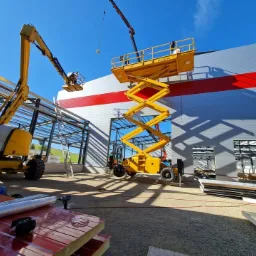 Dwie żółte platformy podnośnikowe przy budowie hali z metalową konstrukcją, widoczne płyty warstwowe i narzędzia na pierwszym planie, budynek z czerwoną linią na szarej elewacji.