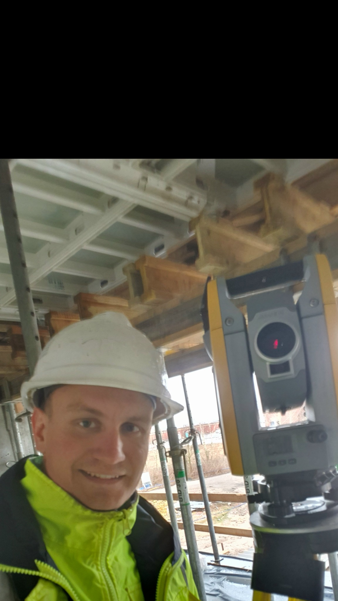 Mężczyzna w kasku i jaskrawej kurtce, robiący selfie z teodolitem na budowie, z widocznym szalunkiem stropowym w tle.