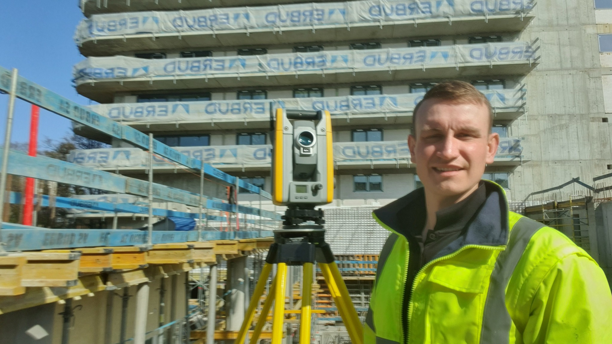 Pracownik w odblaskowej kurtce, obok niego żółty teodolit na trójnogu, w tle surowy budynek w trakcie budowy z zabezpieczeniami na balkonach.