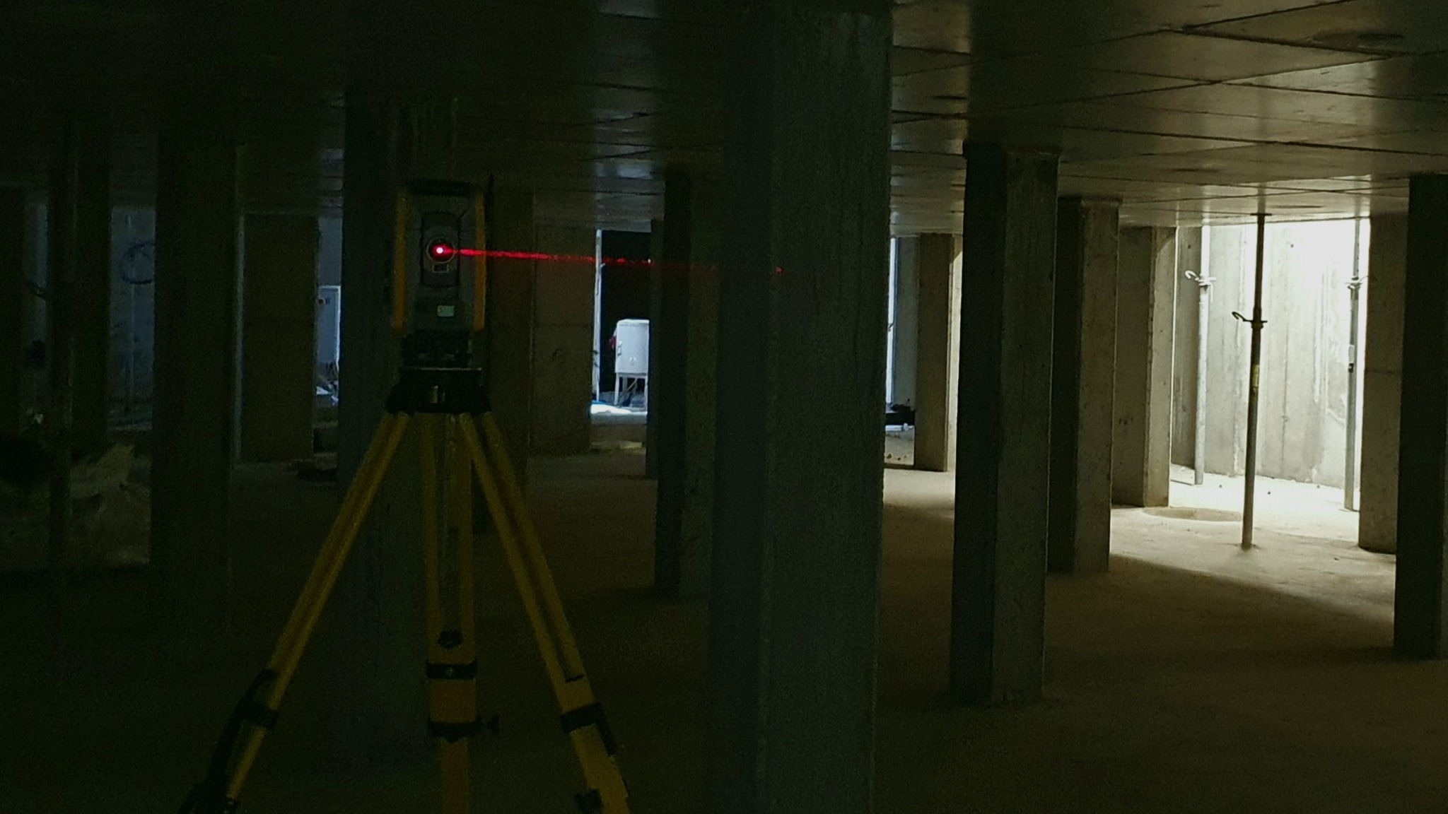 Żółty niwelator laserowy na trójnogu emituje czerwoną wiązkę w surowym wnętrzu budynku w trakcie budowy, widoczne betonowe kolumny i strop.