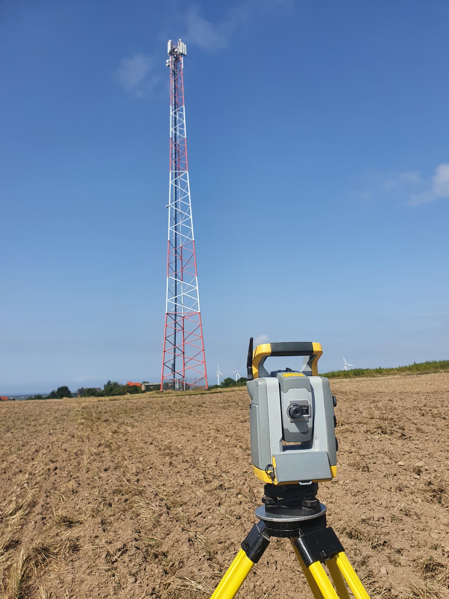 Pomiar geodezyjny wieży telekomunikacyjnej w polu uprawnym, teodolit na statywie z widocznymi turbinami wiatrowymi w tle, błękitne niebo.