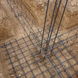 Zbrojenie fundamentu w wykopie, widoczne pionowe pręty i siatka zbrojeniowa, ziemia wokół.