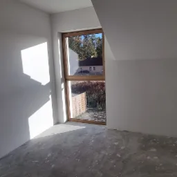 Wnętrze pokoju na poddaszu w stanie deweloperskim z oknem, świeżo po tynkowaniu ścian na biało. Widoczne gniazdka elektryczne i surowa wylewka na podłodze. Promienie słoneczne wpadają przez okno.