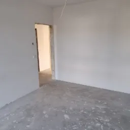 Wnętrze pomieszczenia w stanie deweloperskim z gładkimi, pomalowanymi na biało ścianami, surową wylewką na podłodze, widocznymi gniazdkami elektrycznymi i otwartym przejściem do innego pomieszczenia.