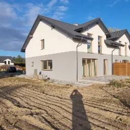 Nowoczesny dom dwukondygnacyjny z szarą dolną i białą górną częścią elewacji, z ciemną dachówką, widoczne okna i drzwi balkonowe z drewnianymi ramami, teren wokół domu częściowo przygotowany...