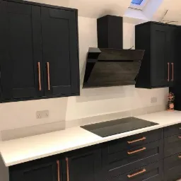 Nowoczesna kuchnia z czarnymi szafkami i białym blatem, miedziane uchwyty, czarna płyta indukcyjna, skośny okap.