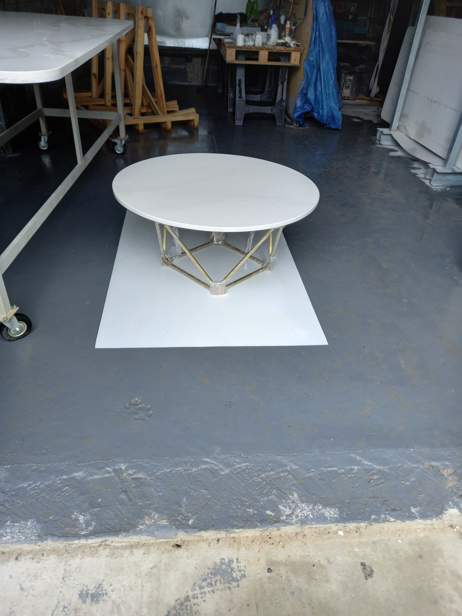 Okrągły blat stołu z jasnego kamienia lub konglomeratu, osadzony na złotym, geometrycznym stelażu, stoi na białej płycie w warsztacie kamieniarskim.